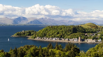 A distant view of Dunollie Wood over the hills behind Oban esplanade with buildings along the shoreline and mountains in the background