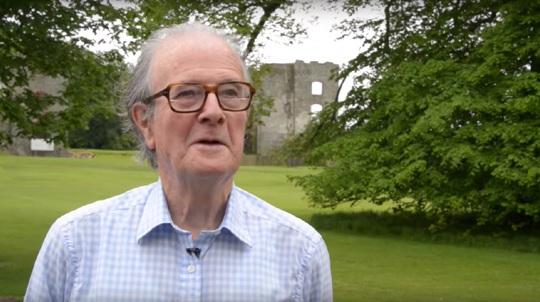 Grey haired man with glasses standing in landscaped grounds talks to camera