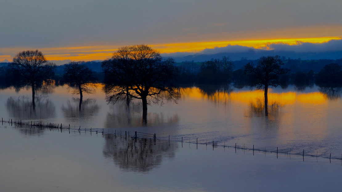 River Severn bursts its banks south of Worcester city centre and water flows over farmland. Clearing skies can be seen in the distance as the sun sets.