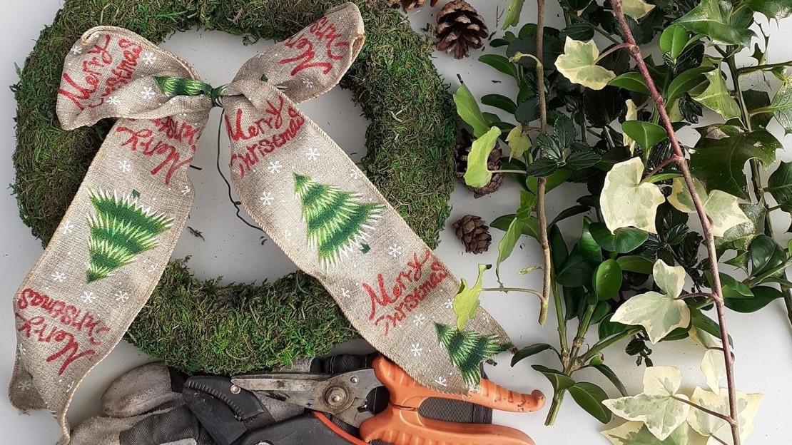 Wreath base, protective gloves, Christmas ribbon, secateurs and foliage arranged on a tabletop