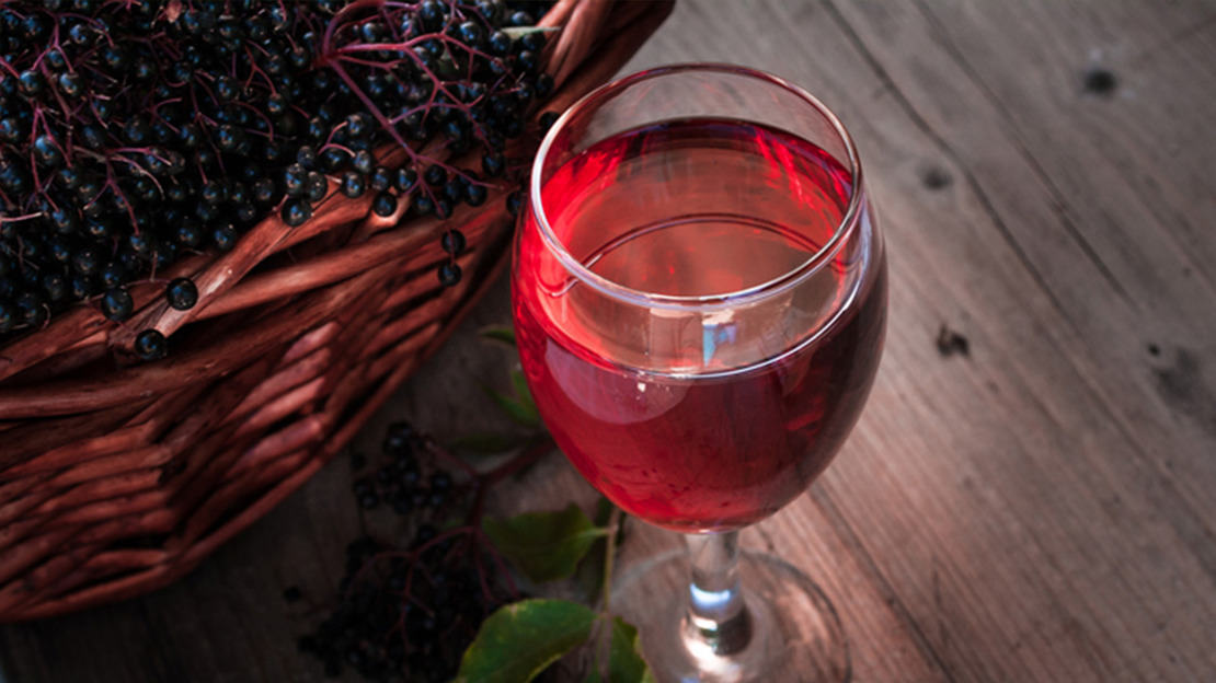 A glass of red elderberry wine next to a basket of elderberries