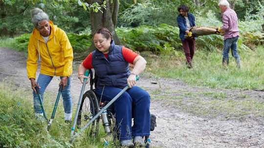 One woman standing and one in a wheelchair use tools to cut plants at ground level