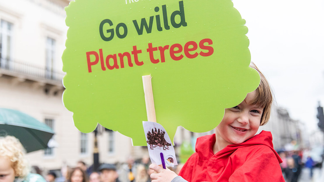 Young child at a protest holding a tree-shaped placard reading 'Go wild, plant trees'.