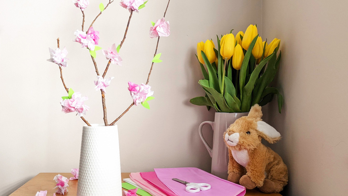 A twig decorated with tissue paper blossom, placed in vase alongside some yellow tulips and a fluffy toy rabbit