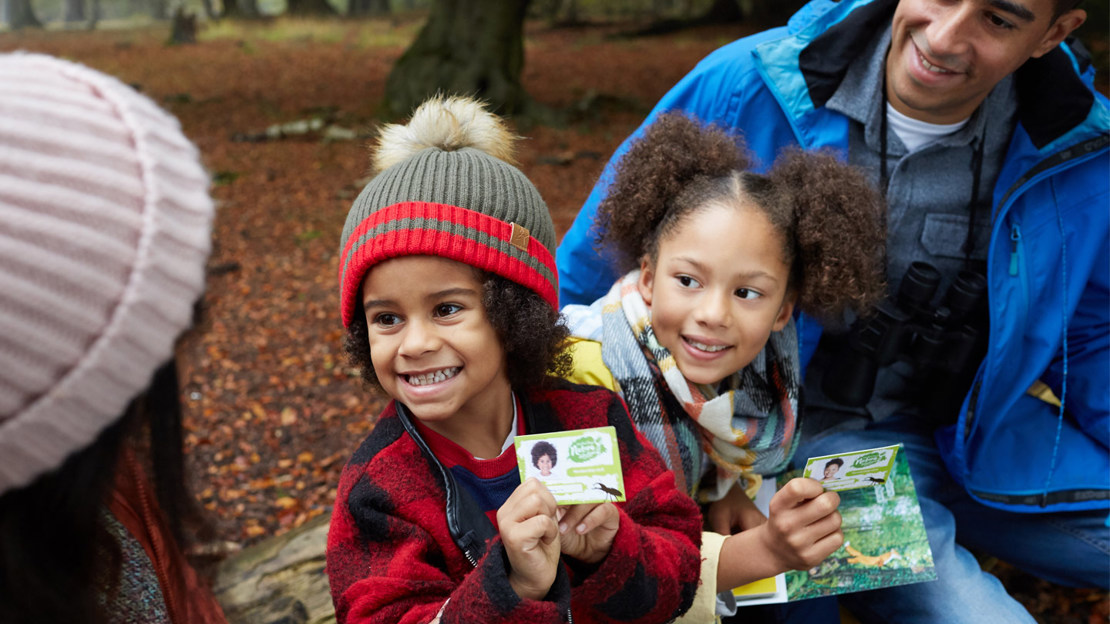 Smiling child holding up a Woodland Trust membership card to their family in woods