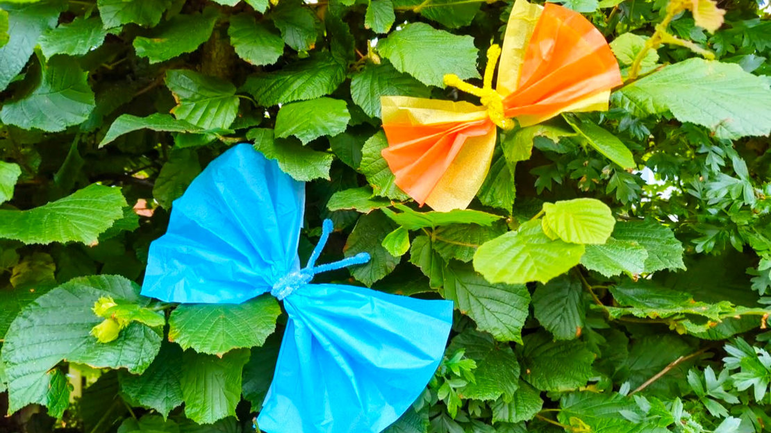 Delicate butterflies made from blue and orange tissue paper, perched on a leafy hazel hedge