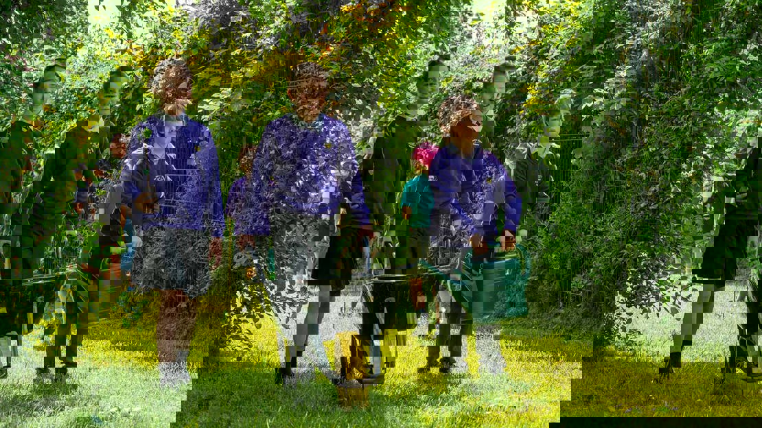 Three children walking through with their schools grounds with a sapling, wheelbarrow and watering can