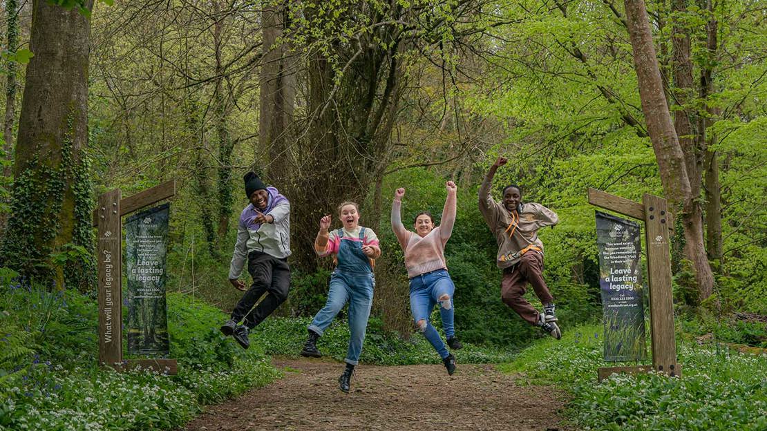 Four young people in a line jumping into the air against a woodland backdrop of trees