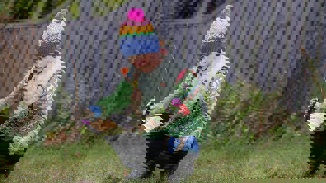 A child in warm clothes crouches in a garden looking at a planting instruction leaflet with a sapling in one hand