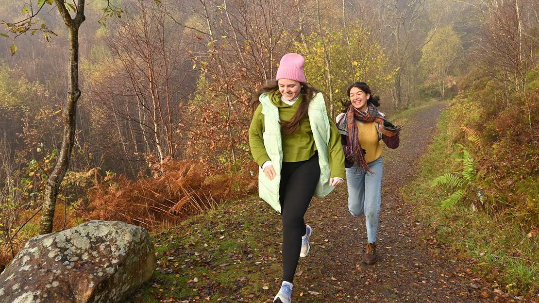 Two young women running along an autumnal woodland path towards camera