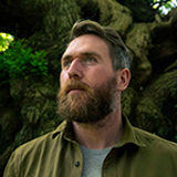Luke Adam Hawker standing in front of a tree trunk Luke Adam Hawker standing in front of a tree trunk