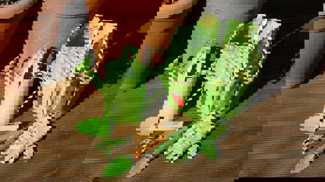 A bright green holdall with child-sized gloves, watering can and garden tools on decking next to terracotta plant pots