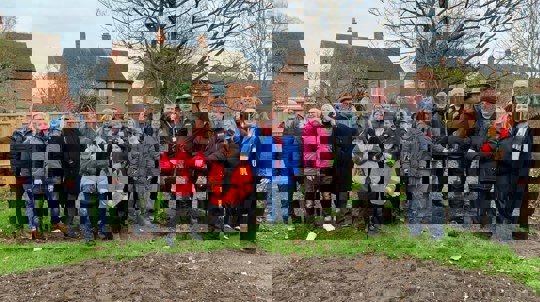 A group of people in winter coats stand in a green space with a mature tree ready to plant.