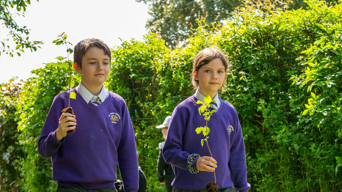 Two pupils in school uniform carrying saplings as they walk past a leafy hedge